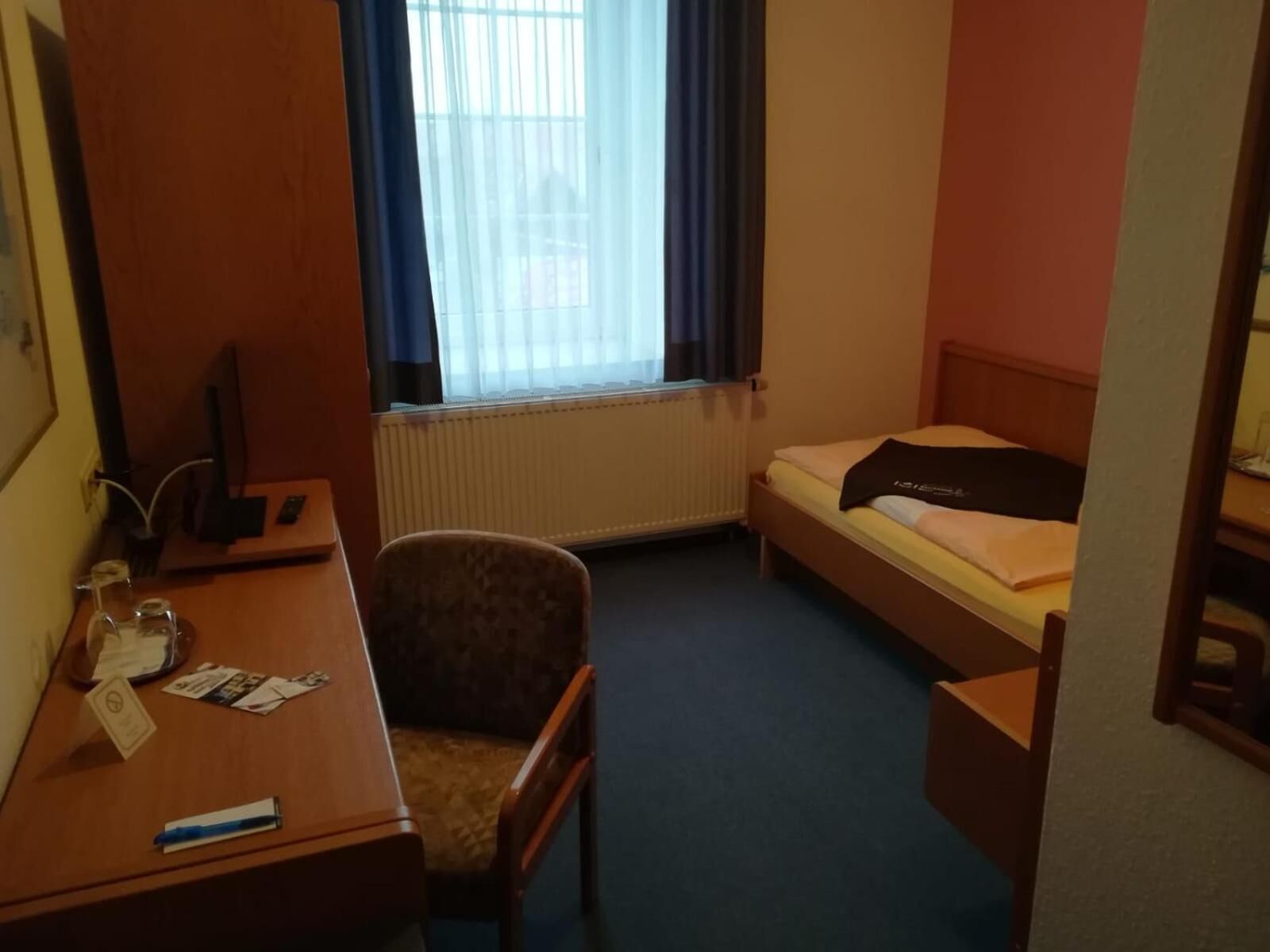 A hotel room with a bed, a desk, a chair, a window with curtains, and a radiator.