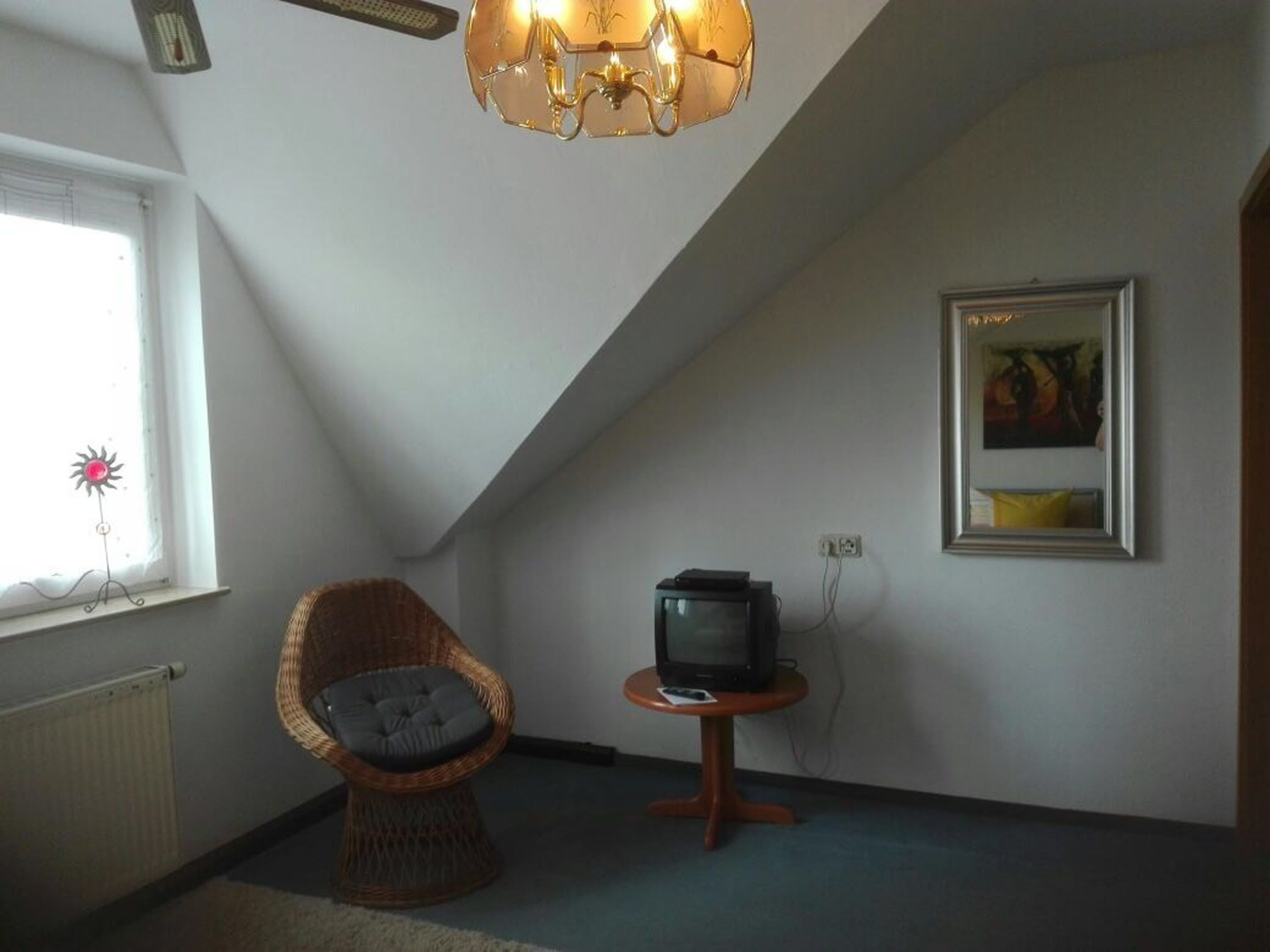 Ein Zimmer mit schrägen Wänden, einem Korbstuhl, einem kleinen Tisch mit Fernseher, einem Heizkörper und einem gerahmten Bild an der Wand.