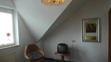 Ein Zimmer mit schrägen Wänden, einem Korbstuhl, einem kleinen Tisch mit Fernseher, einem Heizkörper und einem gerahmten Bild an der Wand.