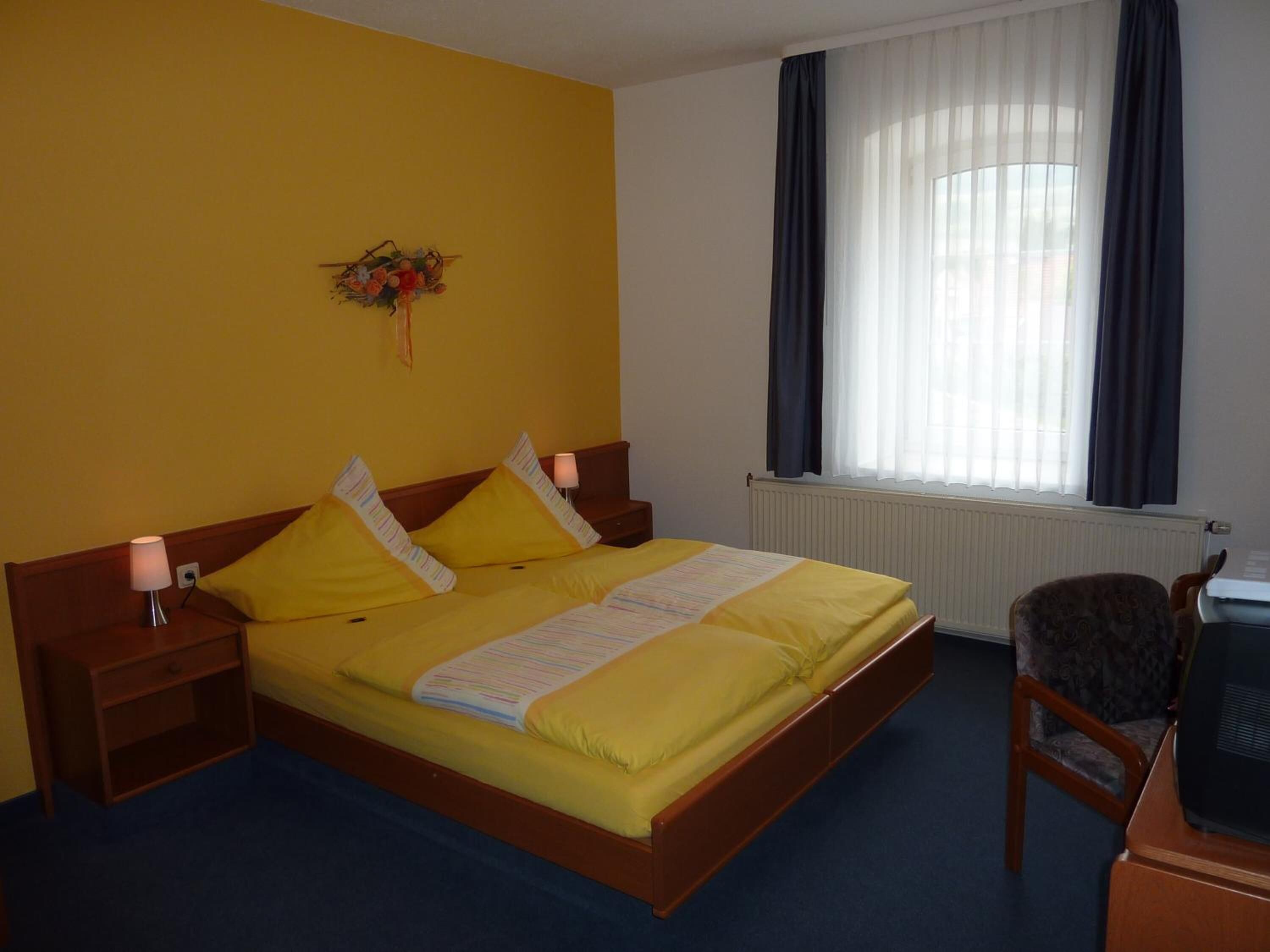 A hotel room with a single bed, a bedside table with a lamp, a chair, a desk with a computer, and a window with curtains.