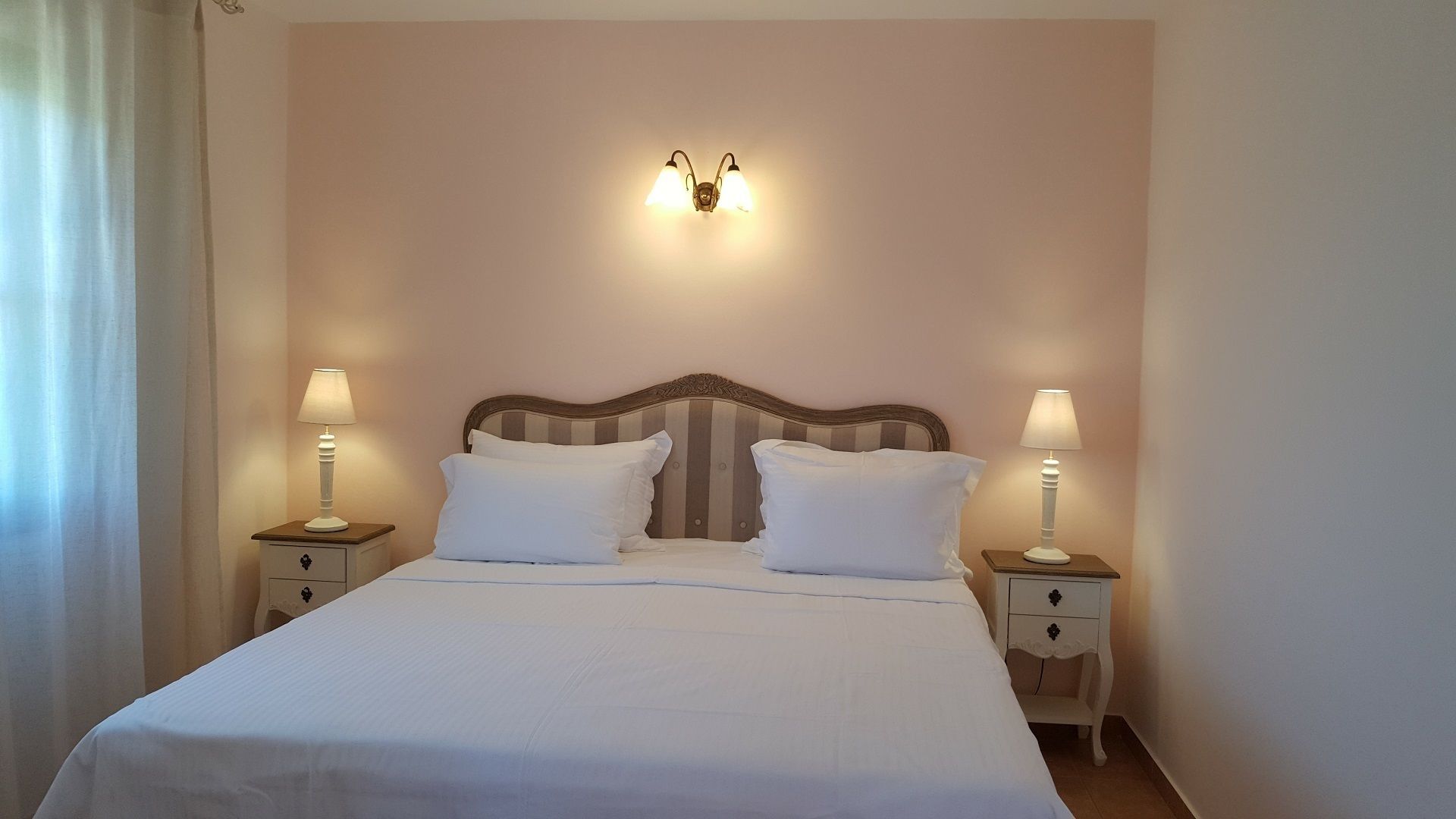 A neatly made bed with white linens and two pillows, flanked by two bedside tables with lamps, in a room with a simple, neutral-colored wall and a decorative light fixture above the bed.
