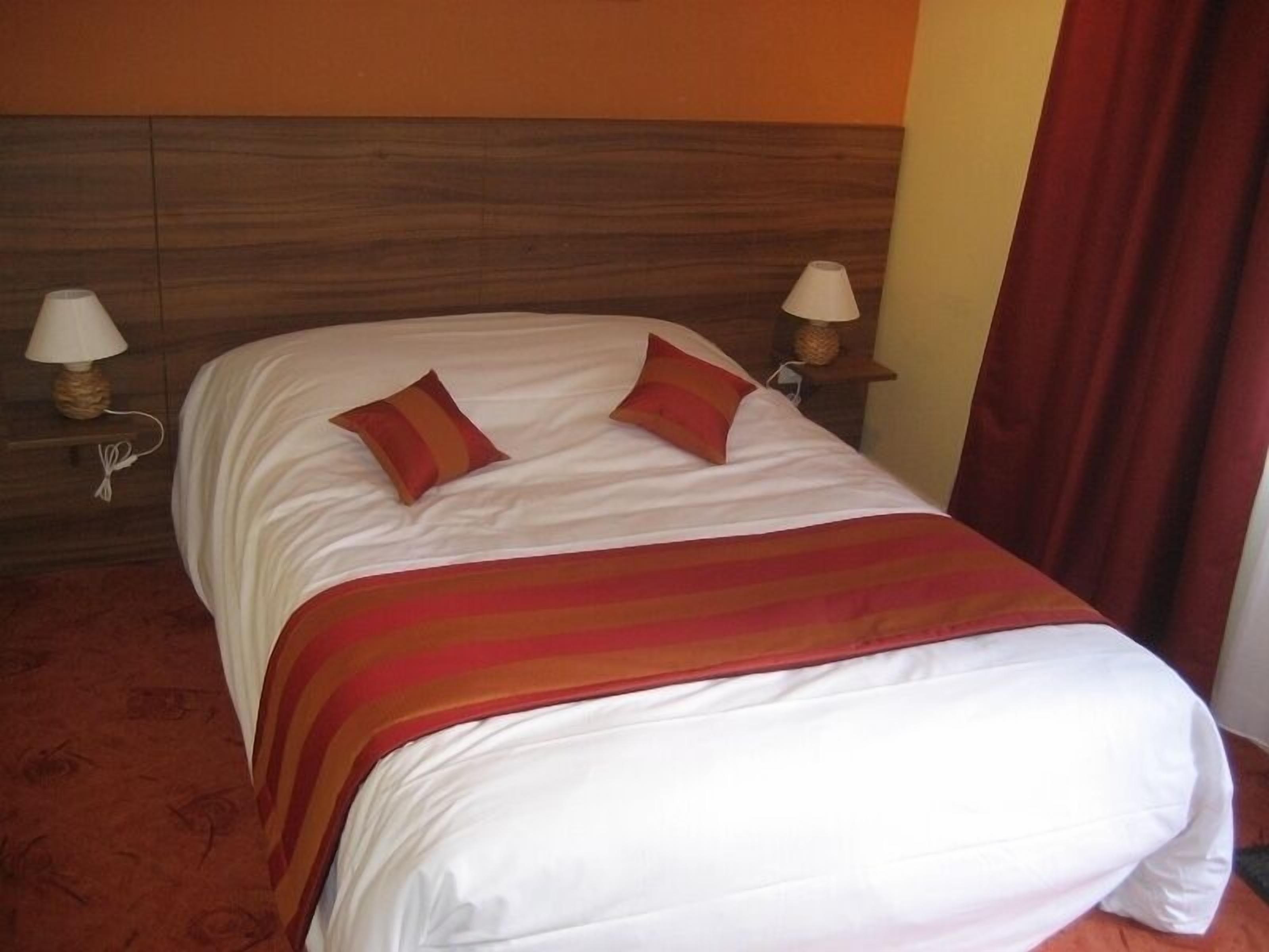 Una cama bien hecha con sábanas blancas y dos almohadas rojas, un cabecero de madera y dos lámparas de noche sobre mesitas de madera.
