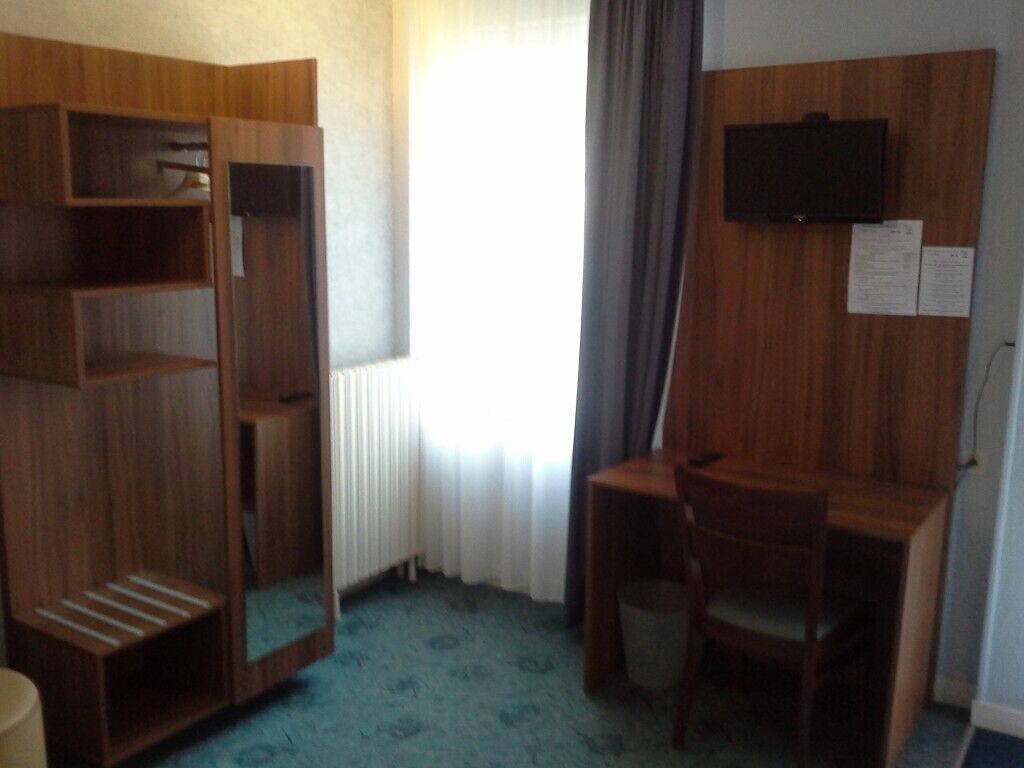 A hotel room with a wooden wardrobe, a desk with a chair, a television mounted on the wall, and a window with curtains.