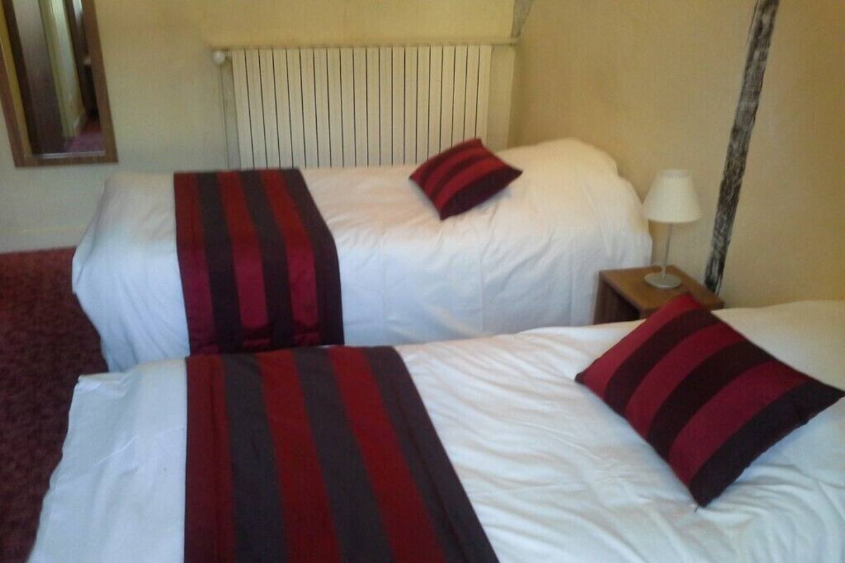 Deux lits avec du linge de lit blanc et des couvertures à rayures rouges et noires, une table de chevet en bois avec une lampe, et un radiateur fixé au mur.
