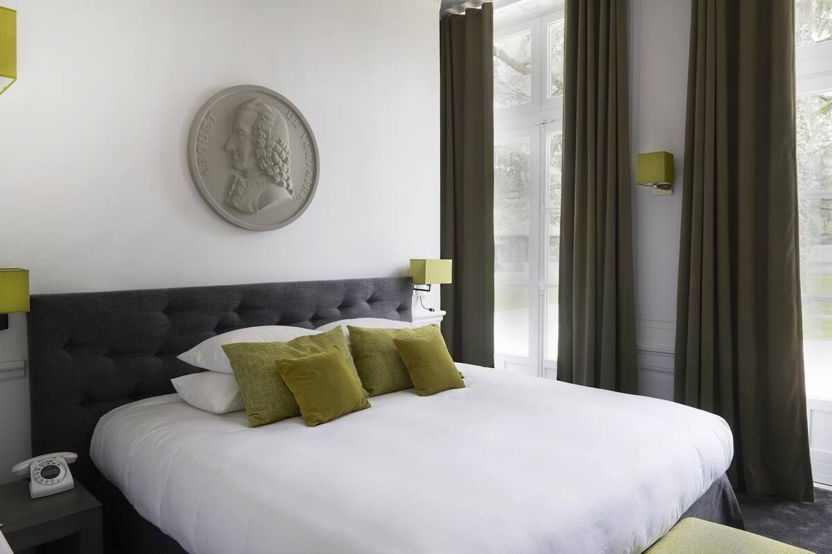 Una cama bien tendida con sábanas blancas y almohadas verdes, un cabecero tapizado oscuro, un teléfono de pared y un plato decorativo sobre la cama.