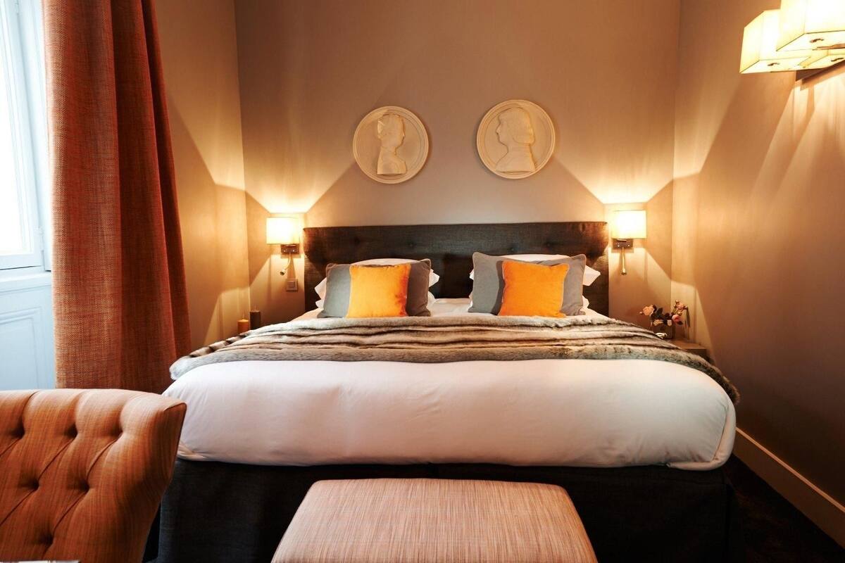 Una habitación de hotel con una cama grande, dos lámparas de pared, un cabecero y un ventanal con cortinas.