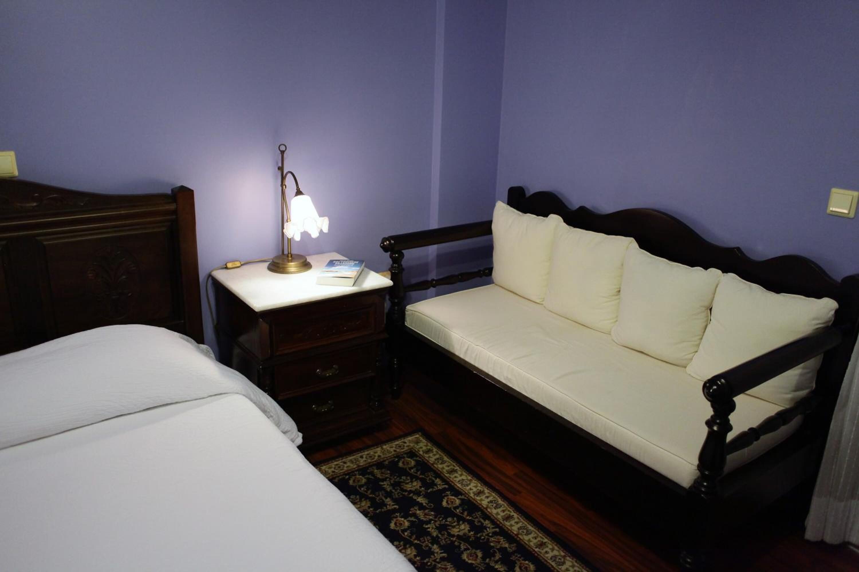 A bedroom with a double bed, a bedside table with a lamp, a wooden bench with cushions, and a patterned rug.