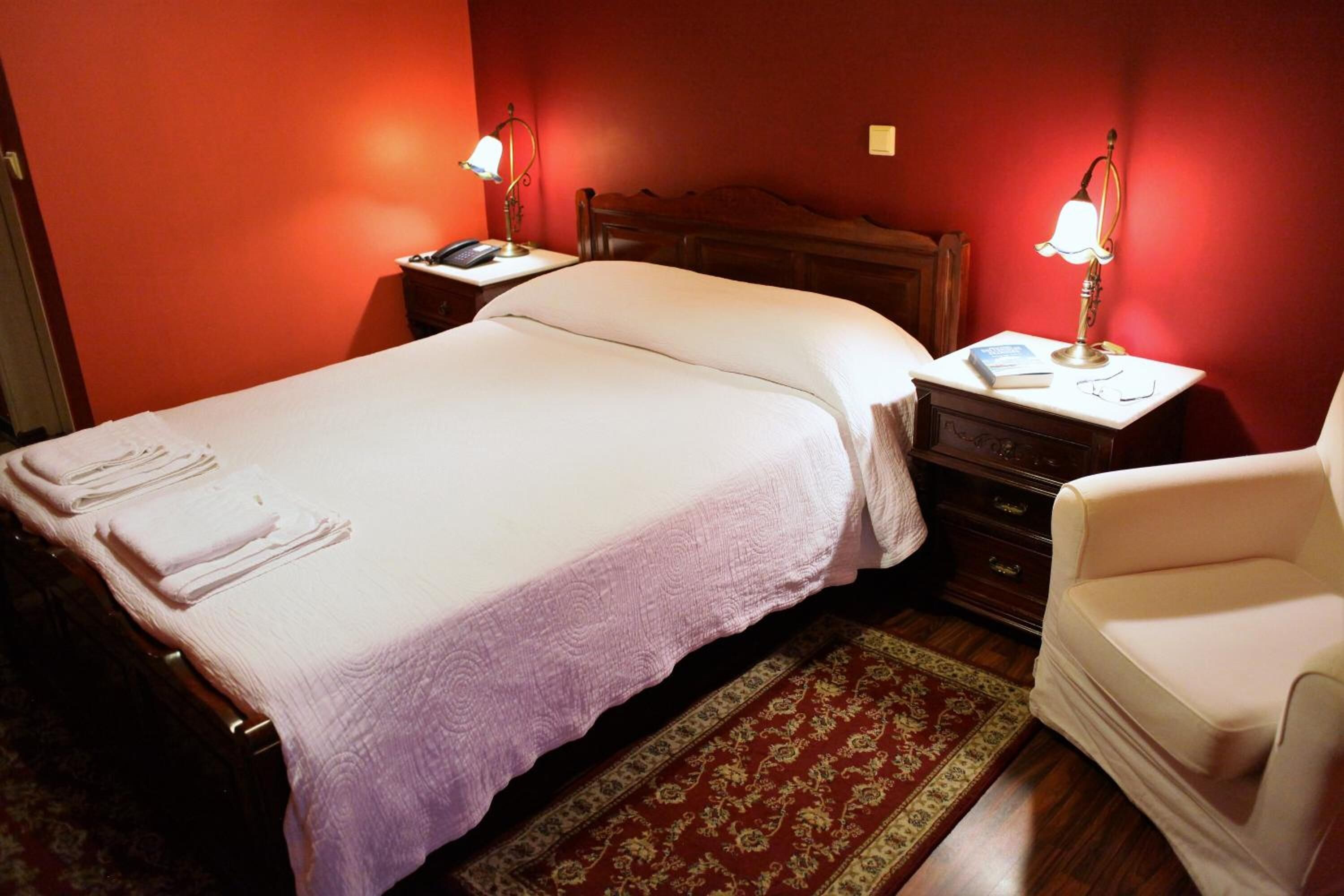 A neatly made bed with a white comforter and pillows, a wooden nightstand with a lamp, a small chair, and a red patterned rug.
