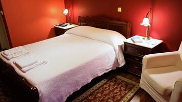 A neatly made bed with a white comforter and pillows, a wooden nightstand with a lamp, a small chair, and a red patterned rug.