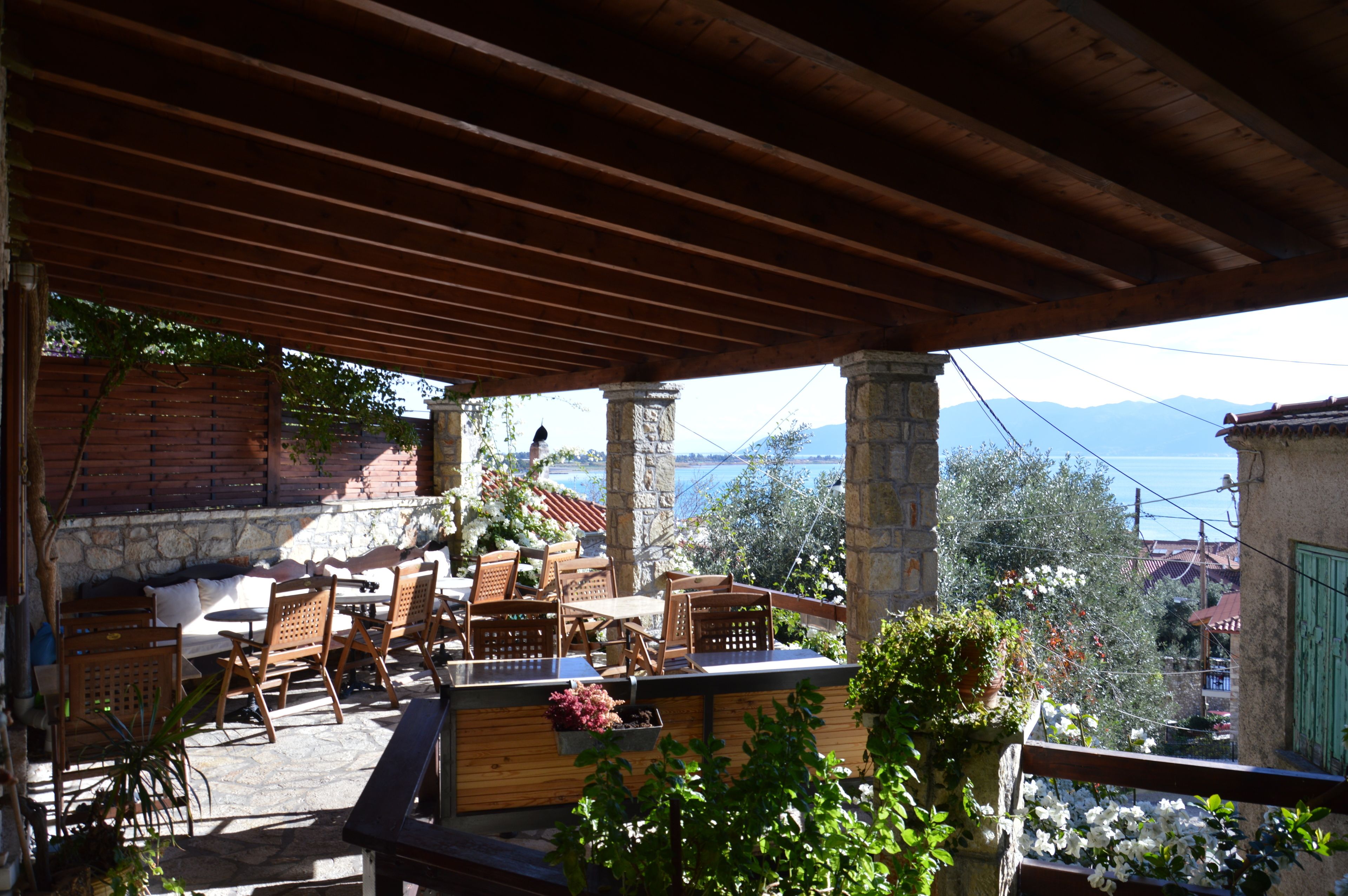 An outdoor patio with wooden furniture and stone pillars, overlooking a scenic view of mountains and a body of water.