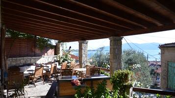 Un patio al aire libre con mobiliario de madera y pilares de piedra, con vistas panorámicas a montañas y una masa de agua.