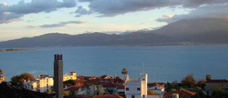 A coastal town with a prominent tower, red-roofed buildings, and a view of the sea and mountains in the background.