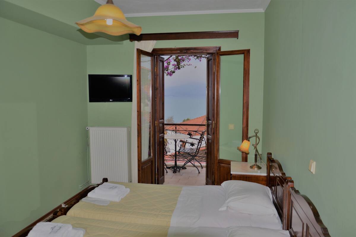 Ein Schlafzimmer mit Bett, Nachttisch, Lampe, einem an der Wand montierten Fernseher und Blick auf einen Balkon mit Geländer.