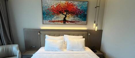 A hotel room with a large bed, a desk, a chair, a painting, and a ceiling-mounted air conditioning unit.