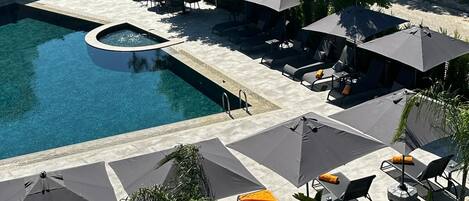 Un área de piscina con sillas de descanso y sombrillas, rodeada de vegetación y edificios.