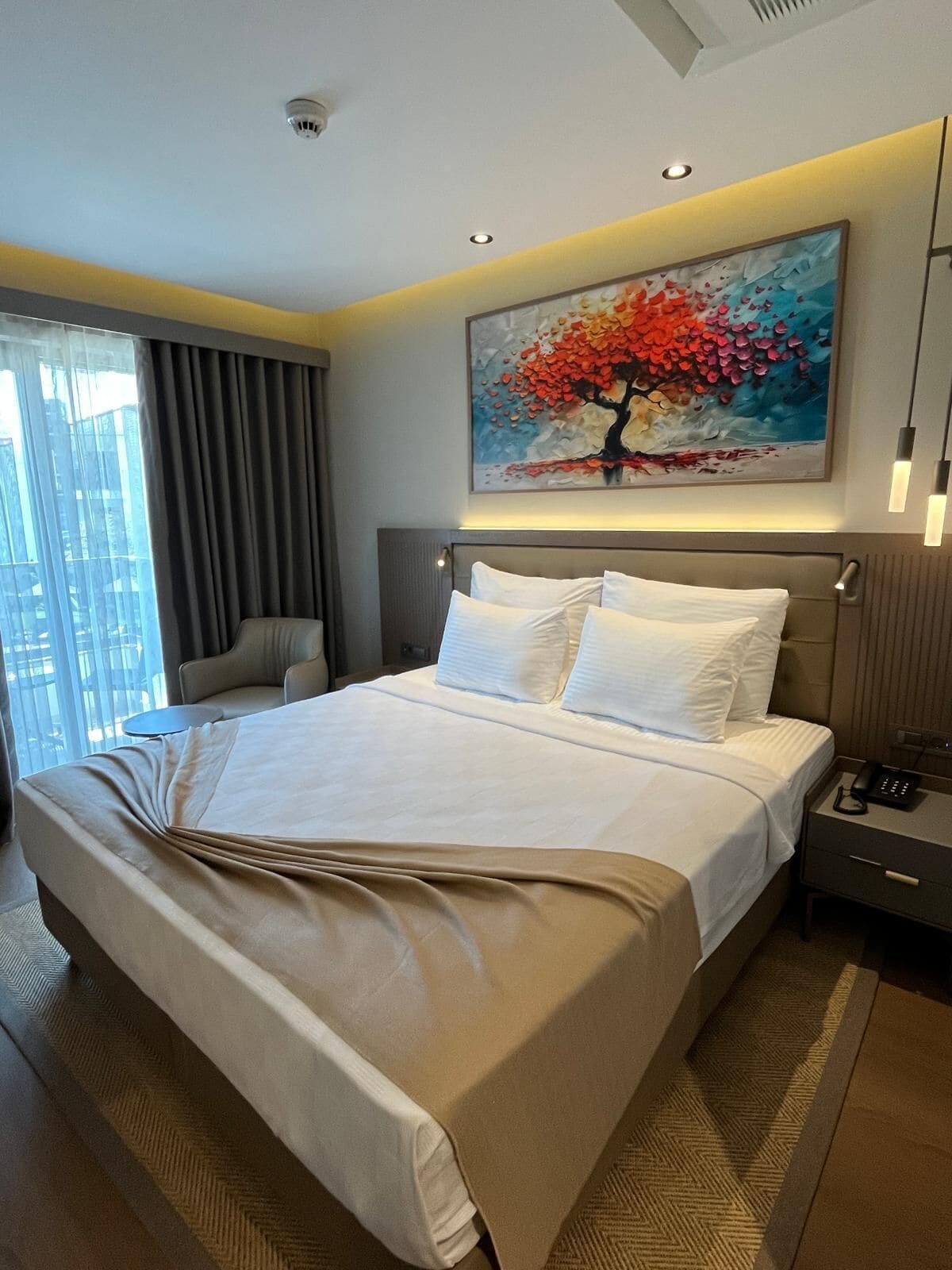 A hotel room with a bed, a bedside table with a phone, a painting of a tree, and a view of buildings through the window.
