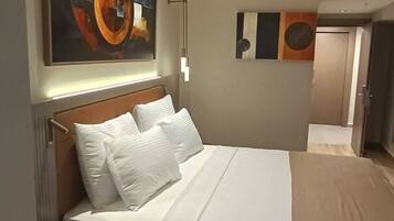 Una habitación de hotel con una cama grande, una mesita de noche con un control remoto, una obra de arte colgada en la pared y una puerta que da a otra habitación.