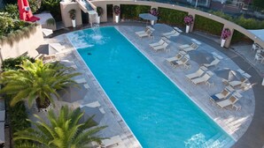 Une piscine rectangulaire à l’eau d’un bleu cristallin, entourée de chaises longues et de parasols, avec des palmiers et des plantes en pot.