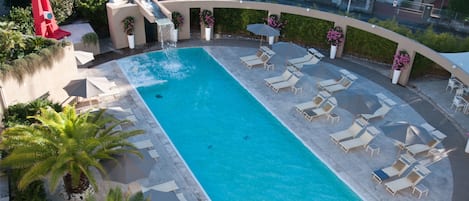 A rectangular pool with clear blue water, surrounded by lounge chairs and umbrellas, with palm trees and potted plants.