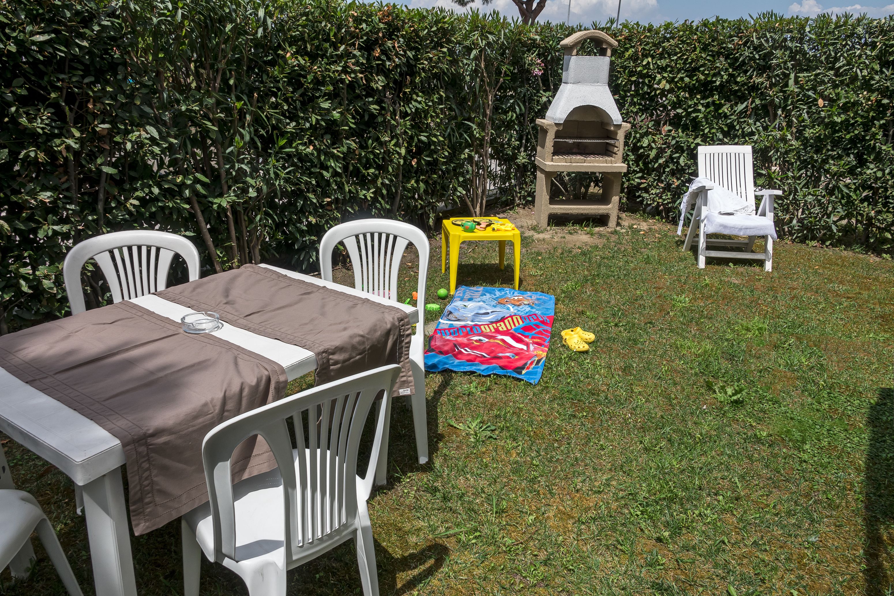 Un patio al aire libre con una mesa preparada para dos, rodeado de sillas de plástico blanco. Se observa una parrilla de barbacoa al fondo, y un taburete amarillo está colocado cerca de la mesa.