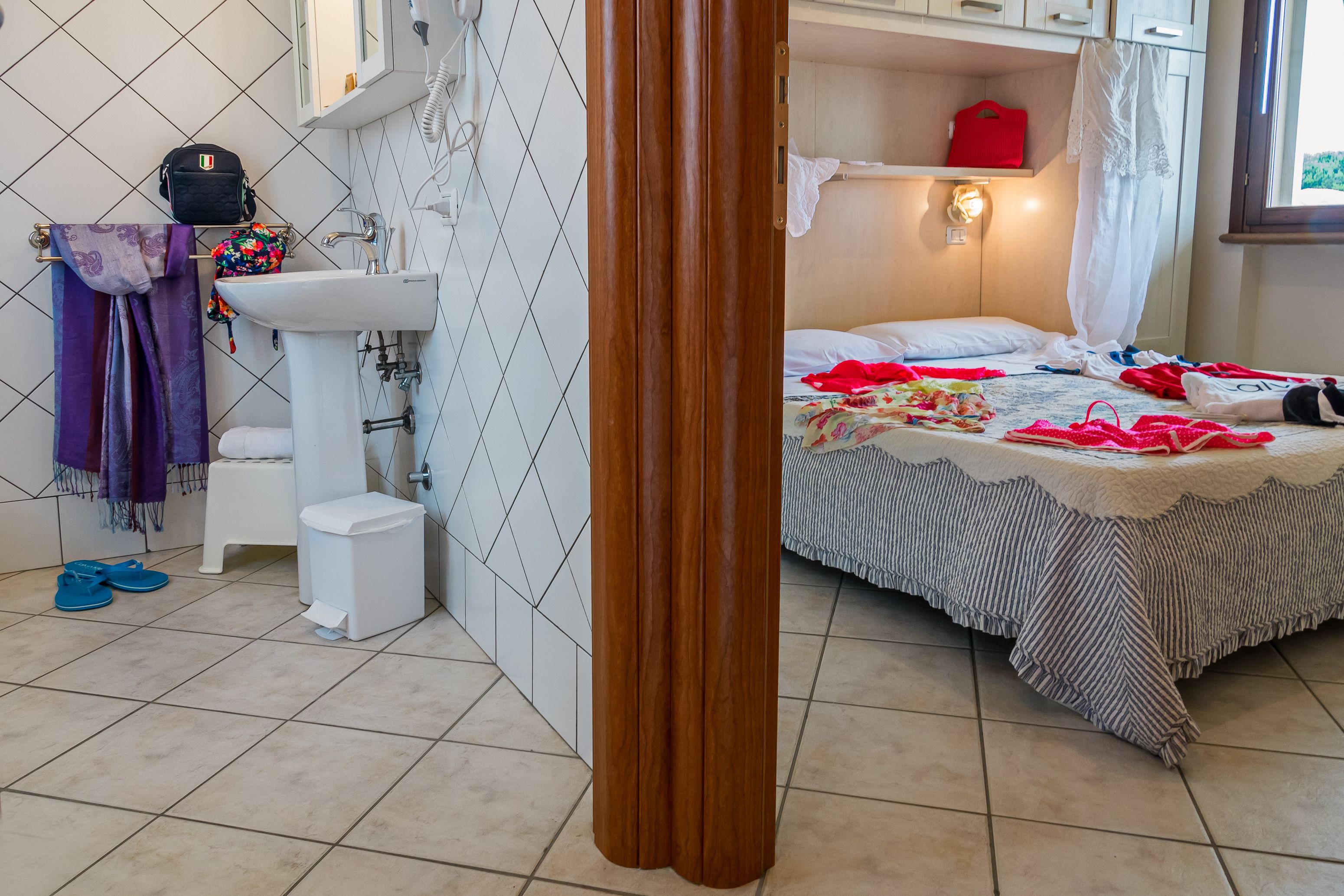A bathroom with a sink, toilet, and a shower area. There is a bed with a striped blanket and a red bag on it.