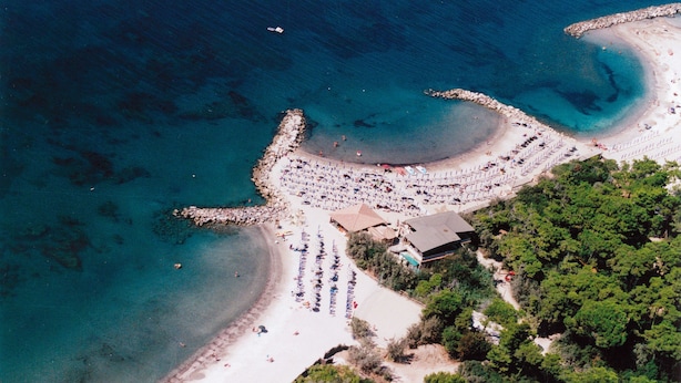 Vue aérienne d’une plage avec une digue rocheuse, une rangée de parasols et un bâtiment doté d’un toit en pente.