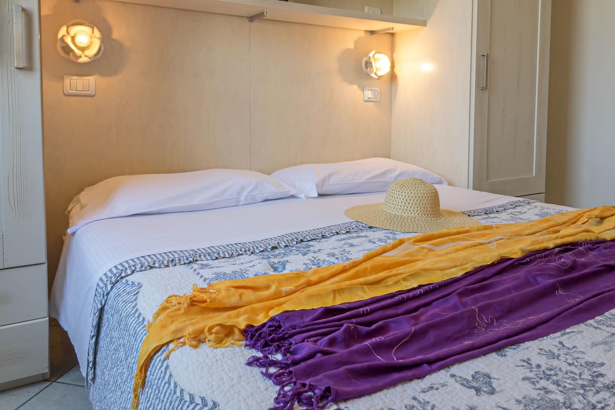 A neatly made bed with white linens, a woven hat, and a colorful blanket.