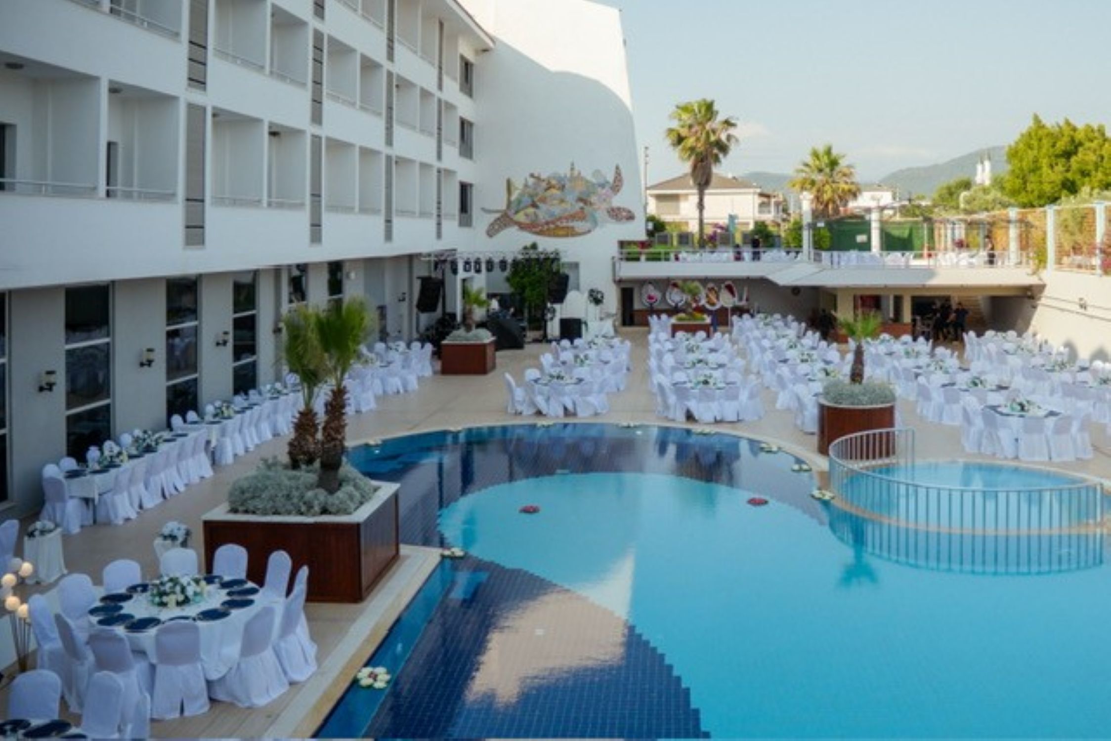 Zona junto a la piscina con mesas y sillas redondas, una piscina grande y un edificio con balcones.