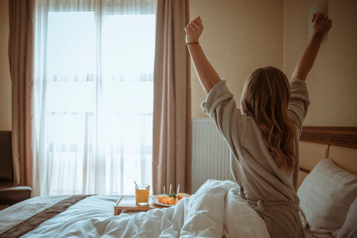 Une chambre d’hôtel avec un lit, une table de chevet sur laquelle se trouve un verre de jus d’orange et une assiette de fruits, et une vue sur un immeuble par la fenêtre.