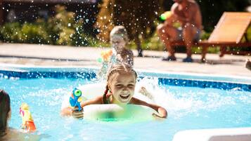 Una piscina con un niño jugando en el agua, rodeada de sillas de descanso y una persona al fondo.