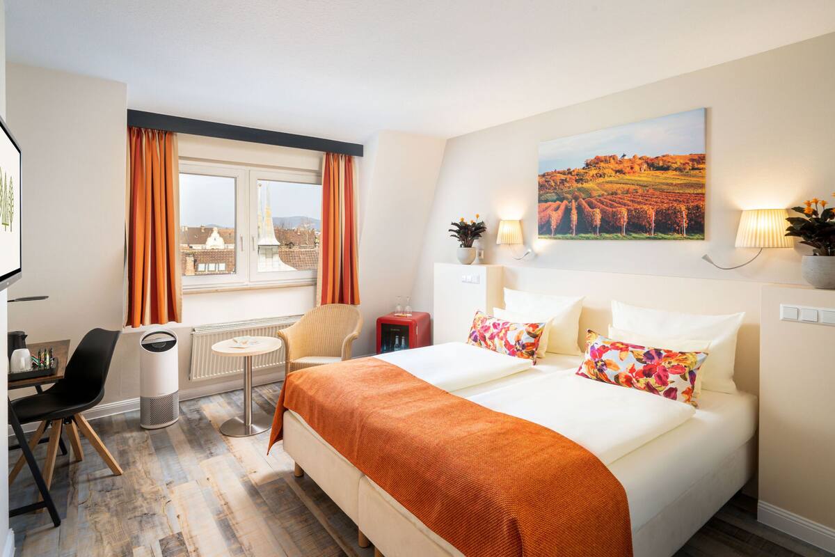 A hotel room with a bed, a desk, a chair, a radiator, and a window with curtains.