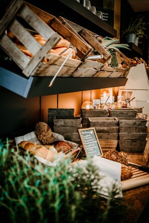 A bakery display with various baked goods in wooden crates and trays, a chalkboard sign listing items, and a warm interior lighting. - Parkhotel Landau (Landau in der Pfalz)