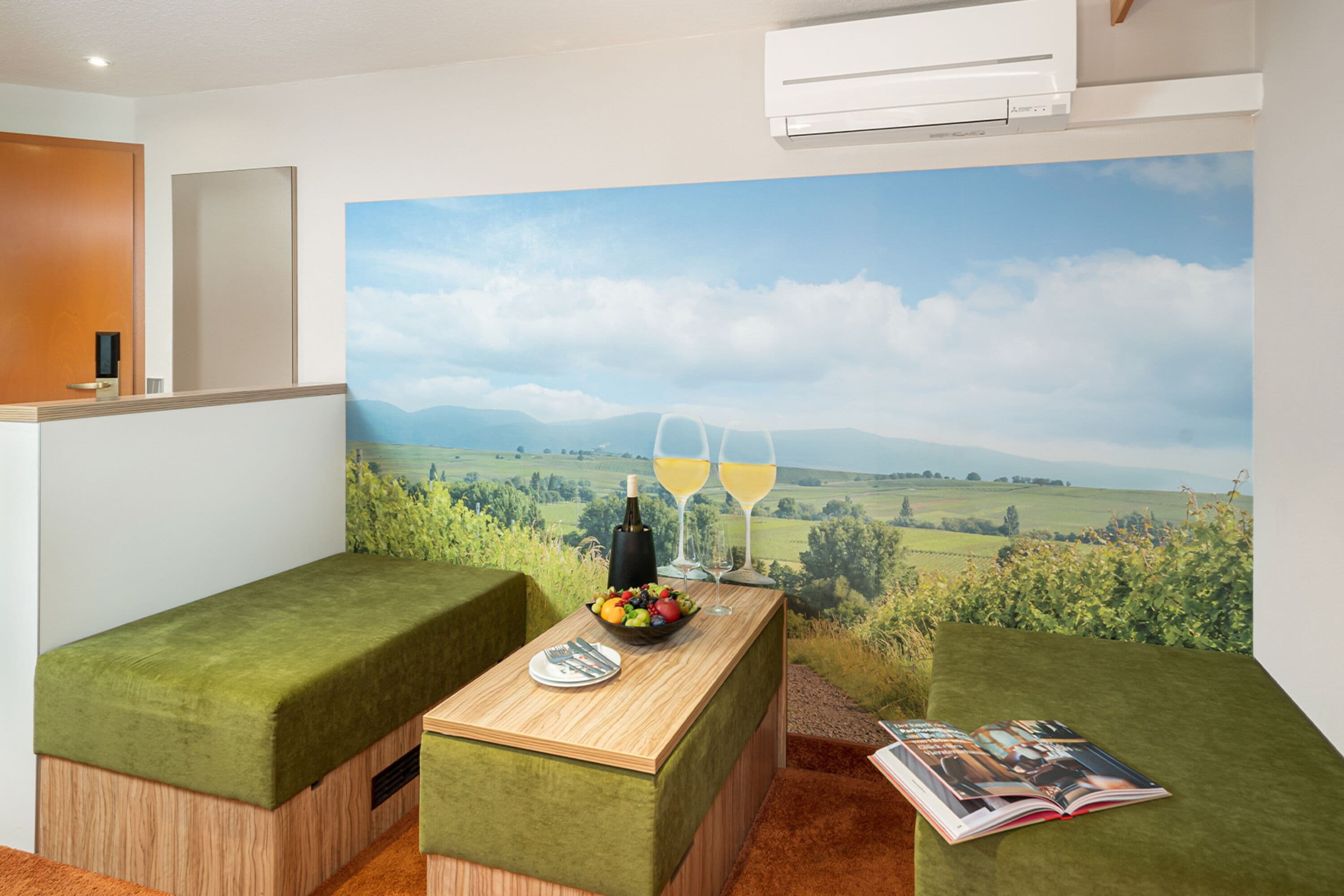 A room with a green upholstered bench, a wooden table with a fruit bowl and wine glasses, and a large landscape mural on the wall.
