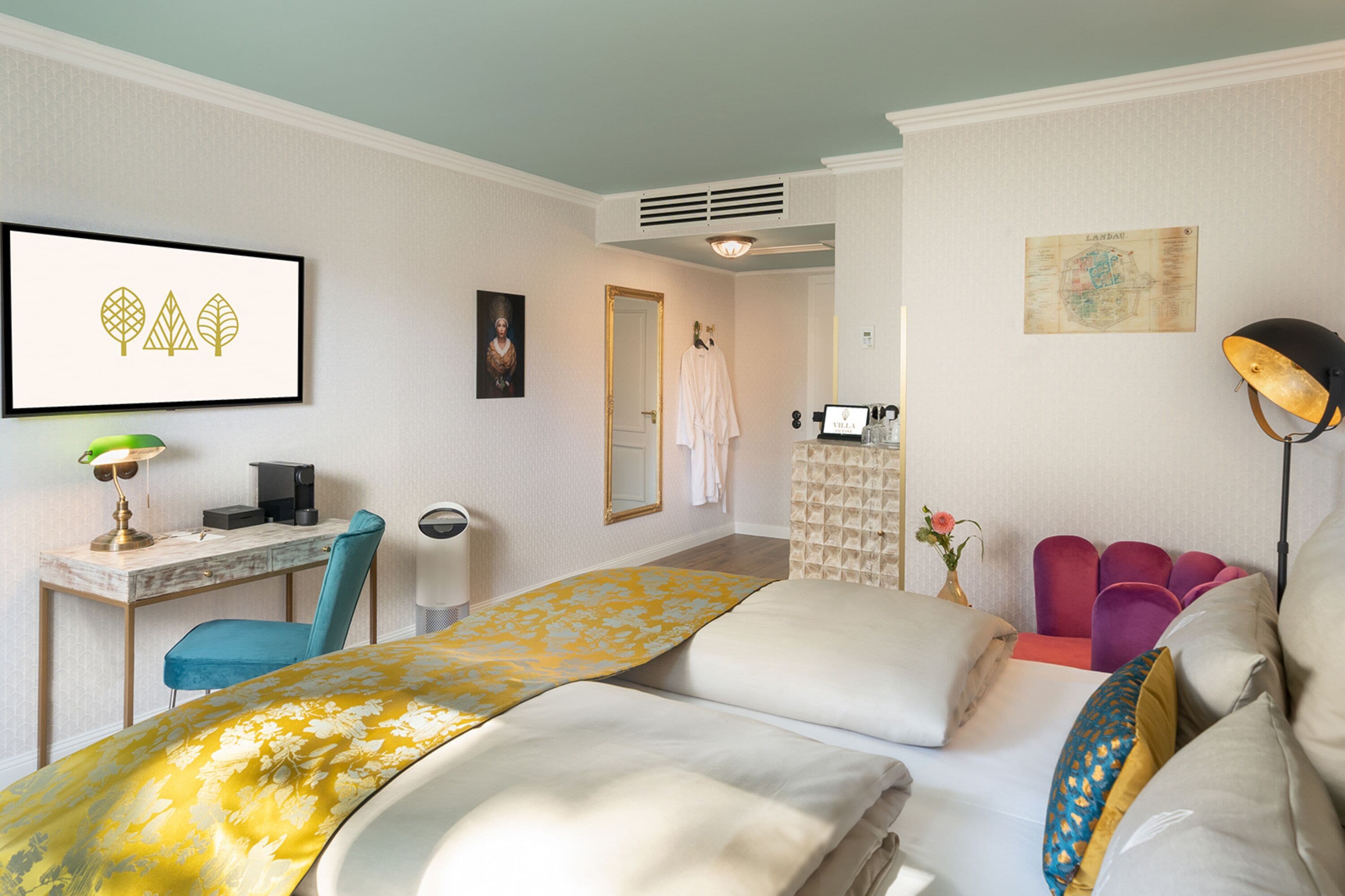 A hotel room with a bed, a desk with a computer, a chair, a lamp, and a map on the wall.