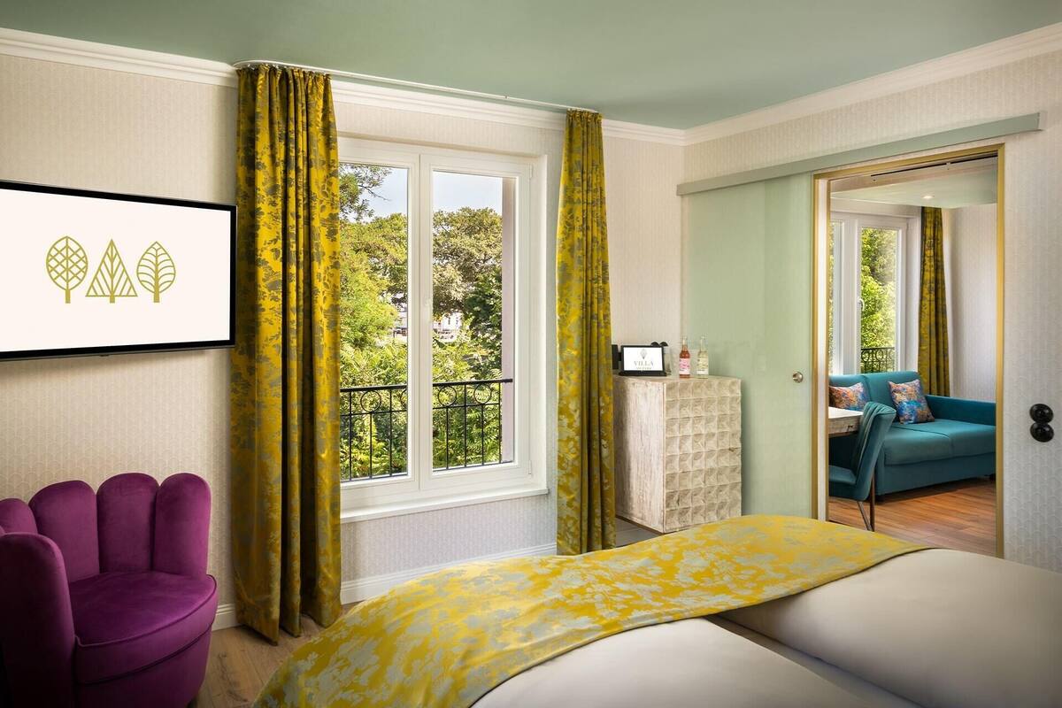 A bedroom with a bed, a purple chair, a window with curtains, a small cabinet, and a view of a balcony with a sofa.