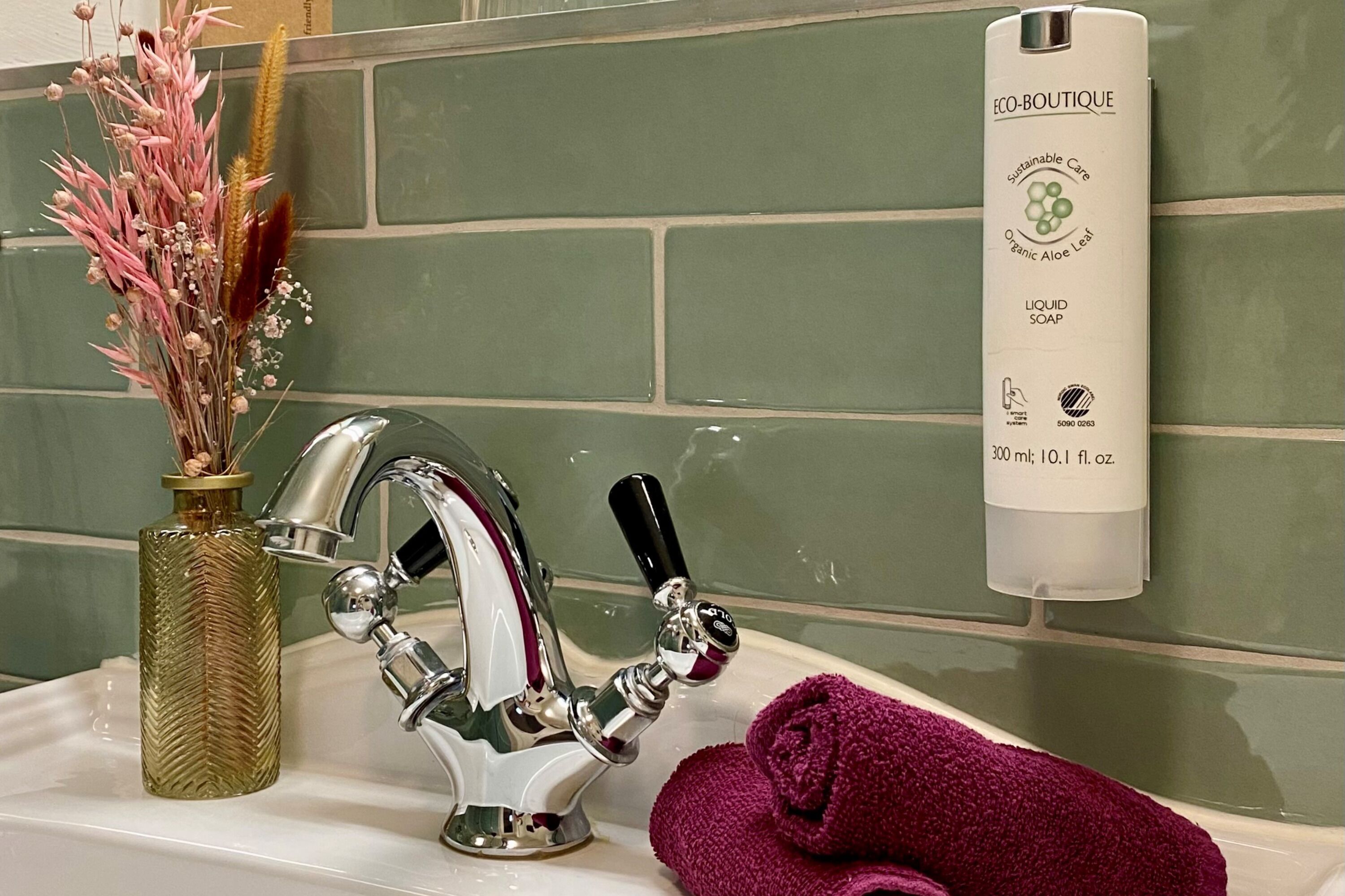 Ein Waschtisch mit einem modernen Wasserhahn, einer Seifenspenderflasche und einer dekorativen Vase mit Trockenblumen.
