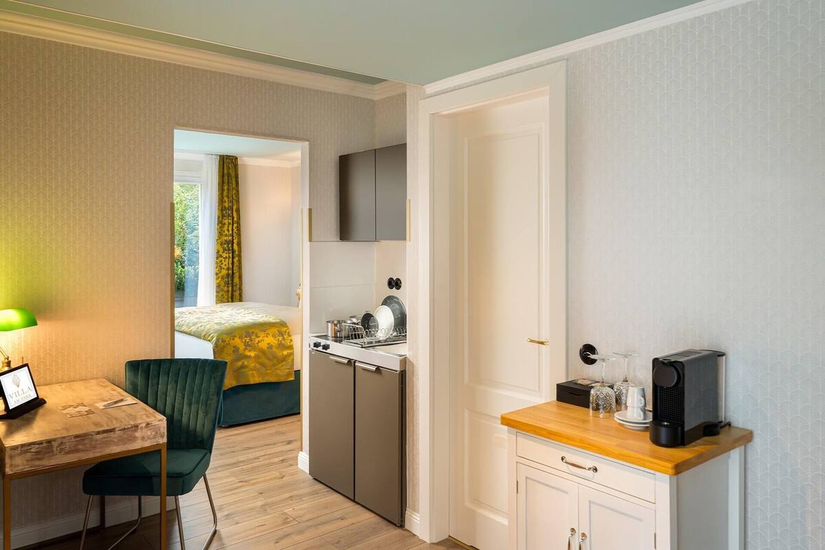 A compact hotel room with a bed, a desk with a chair, a kitchenette, and a TV mounted on the wall.