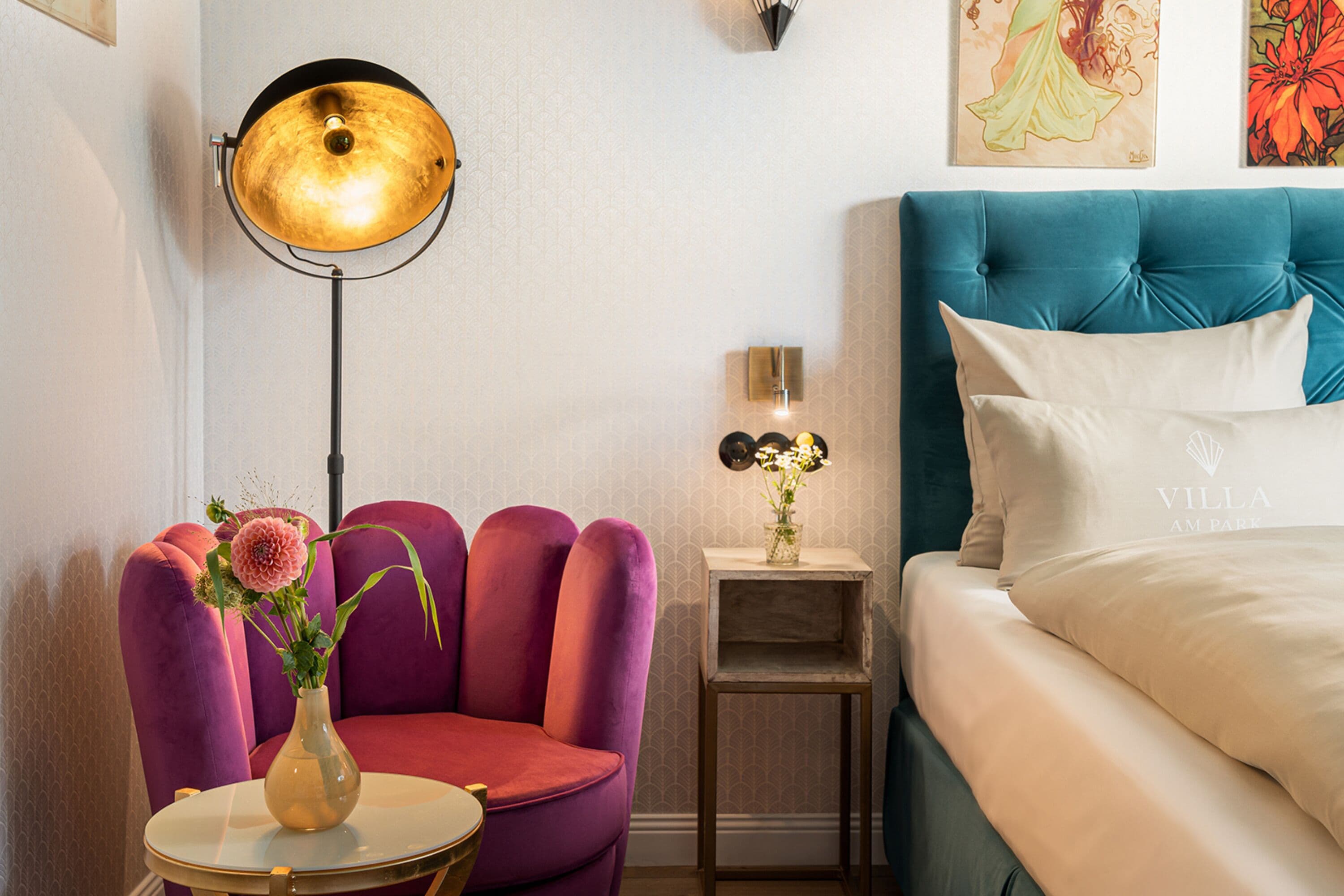 Habitación de hotel con cabecera tapizada en color verde azulado, cama con ropa blanca, una silla rosa con un florero, una mesa redonda con un jarrón dorado y una lámpara de pie.
