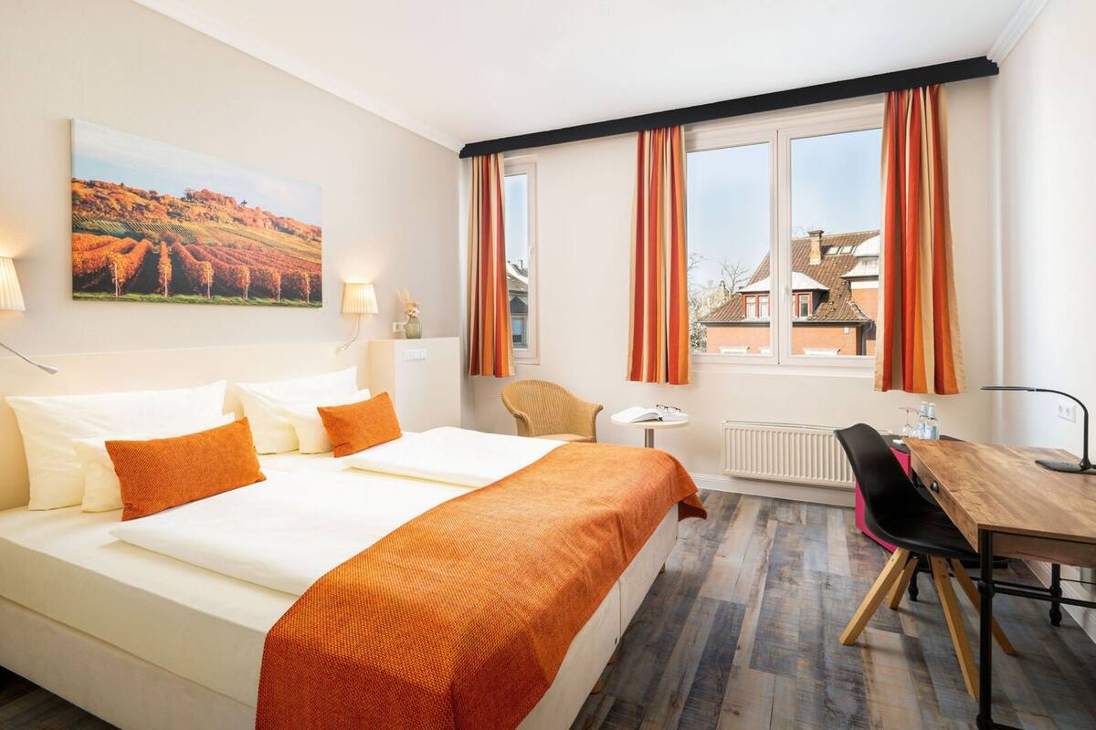 A hotel room with a bed, a desk, a chair, a window with curtains, a radiator, and a painting on the wall.