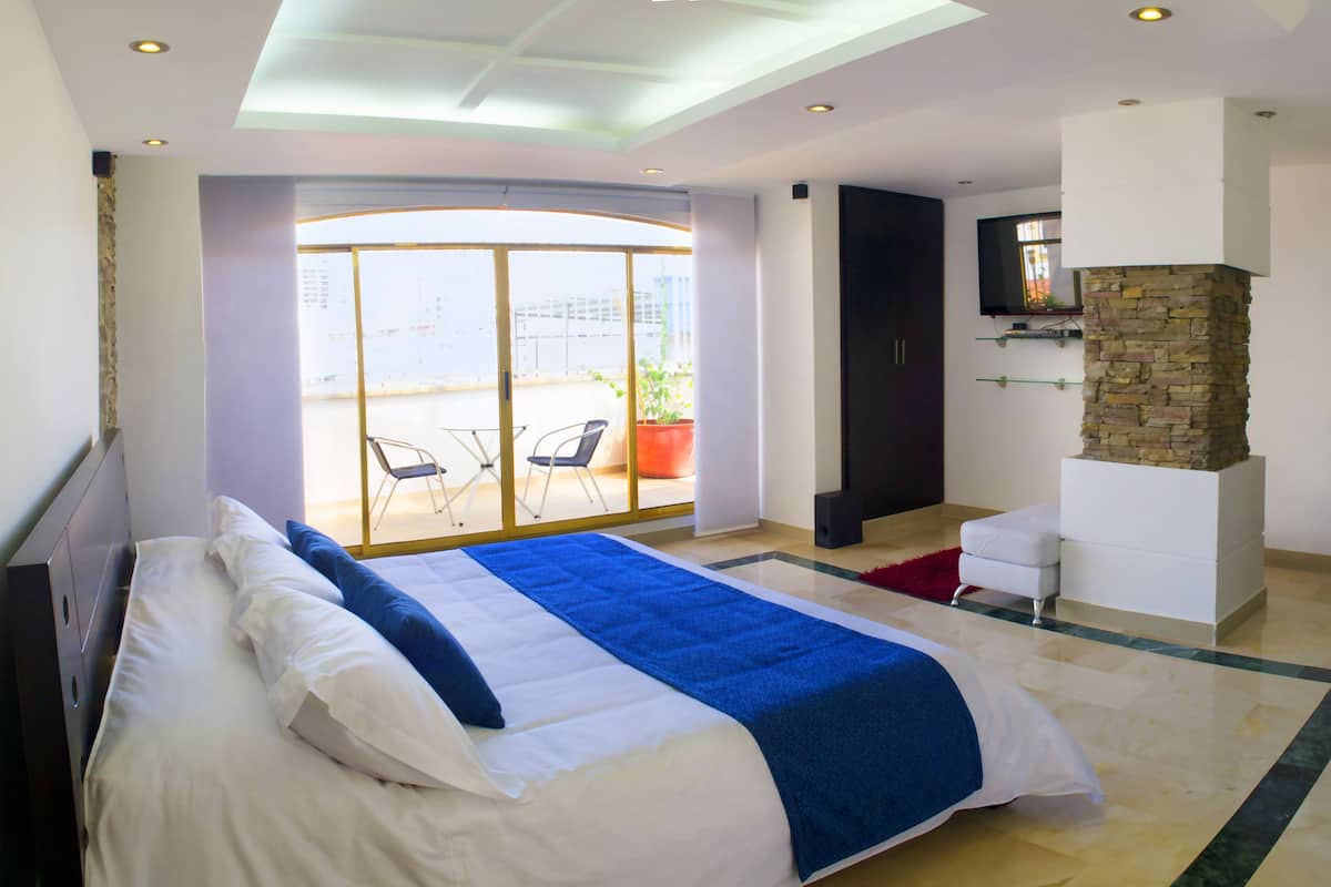 A modern hotel room with a large bed, a stone fireplace, a balcony with a chair, and a TV mounted on the wall.