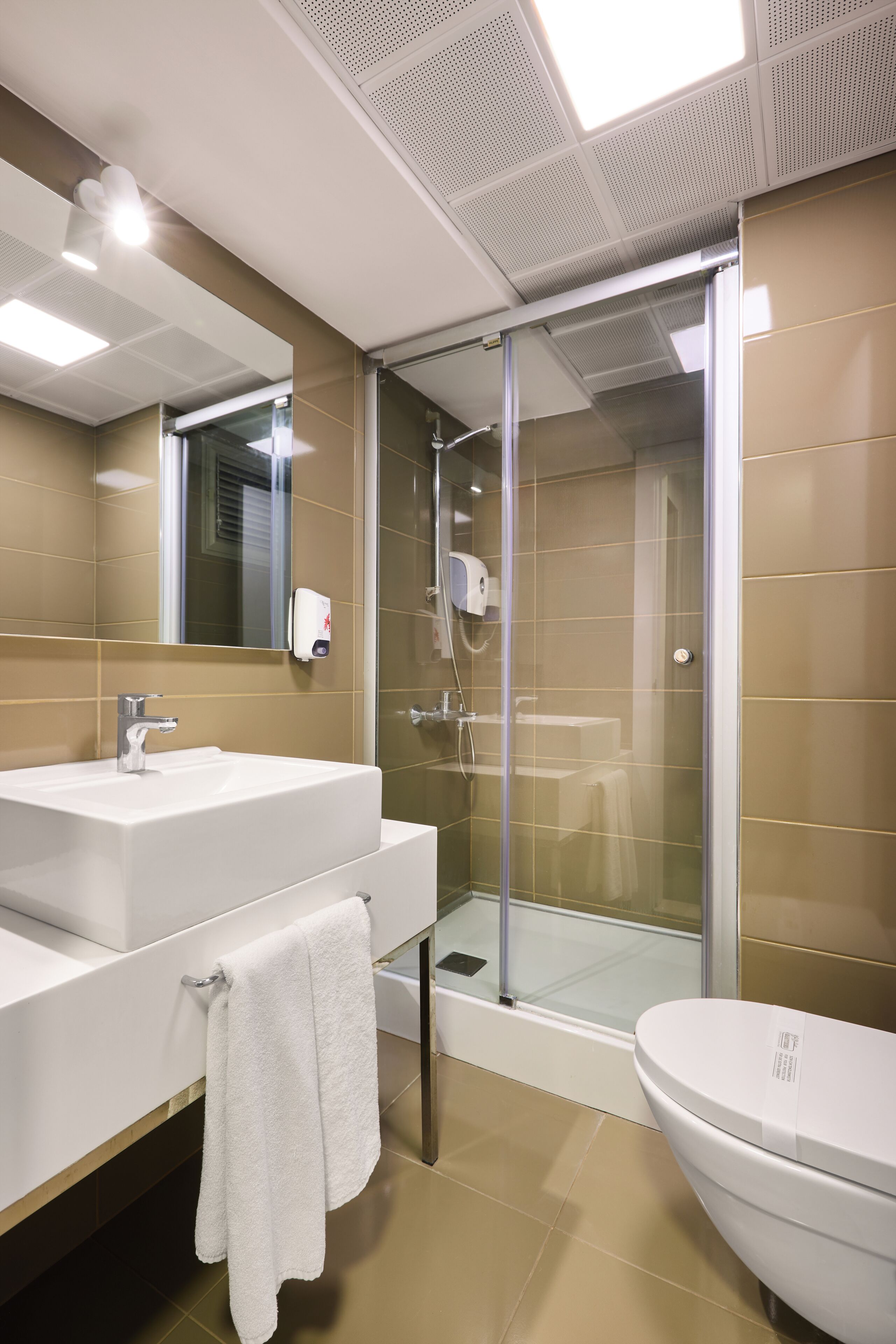 Une salle de bain avec un lavabo blanc, un miroir et des toilettes, une douche en verre et des murs recouverts de carreaux beiges.