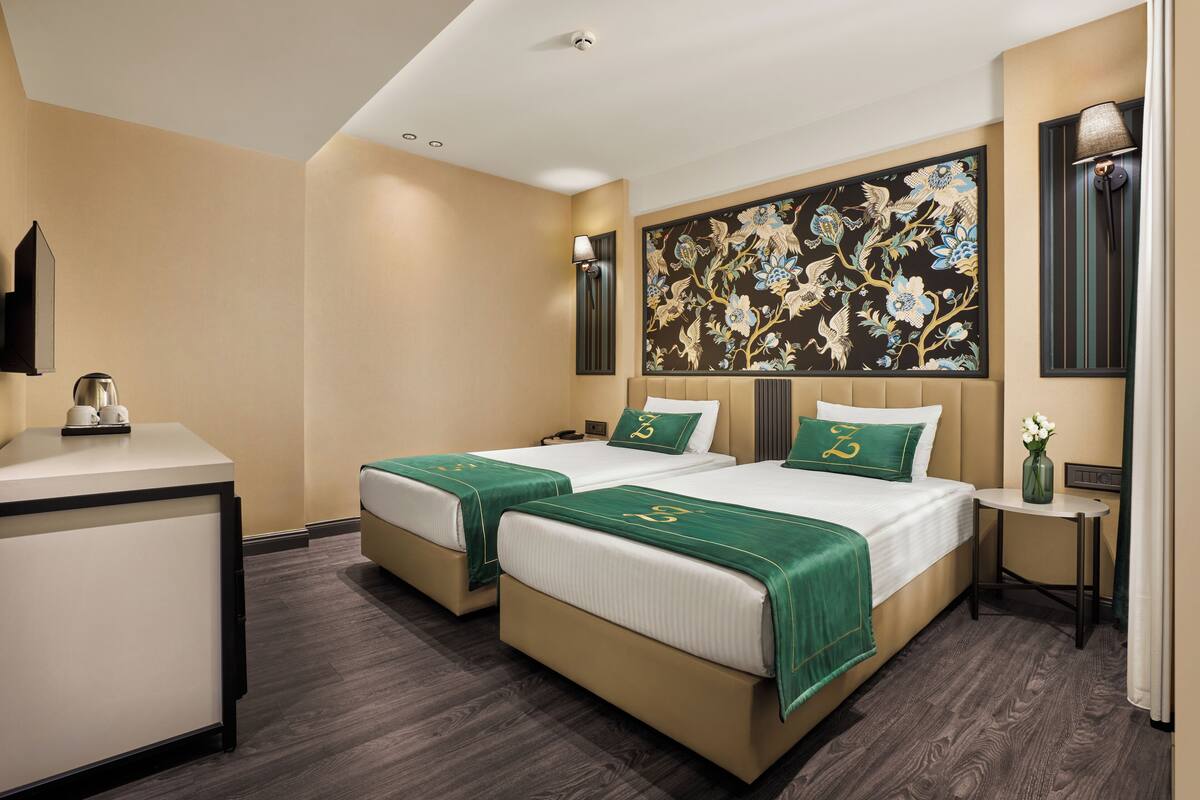 A hotel room with two beds, a desk, a wall-mounted lamp, and a floral artwork.