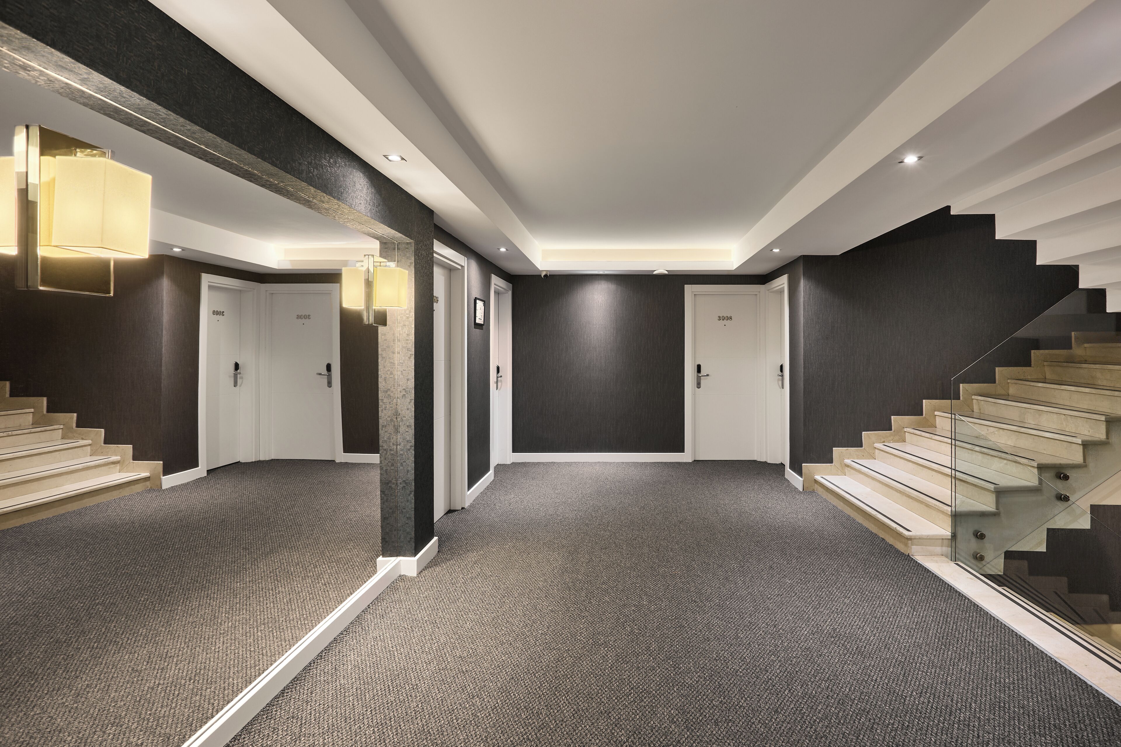 Un pasillo moderno de hotel con pisos alfombrados, una escalera de cristal y una pared oscura.