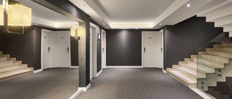 Un pasillo moderno de hotel con pisos alfombrados, una escalera de cristal y una pared oscura.
