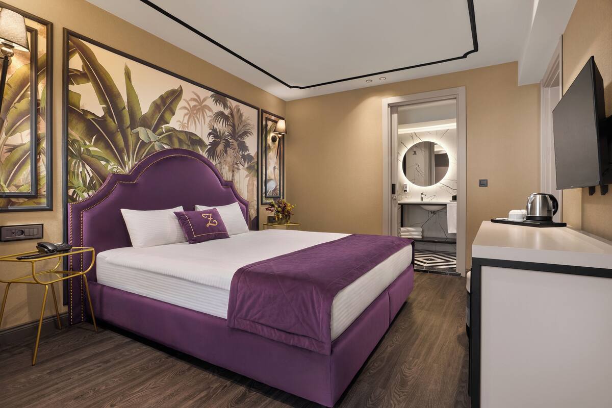A modern hotel room with a large bed, a desk, a mirror, and a wall with tropical artwork.