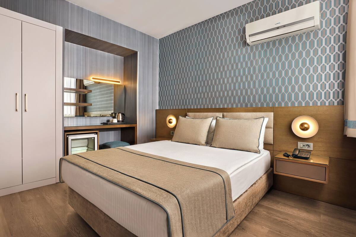 A modern hotel room with a bed, bedside table, nightstand, and a built-in desk with a mirror.