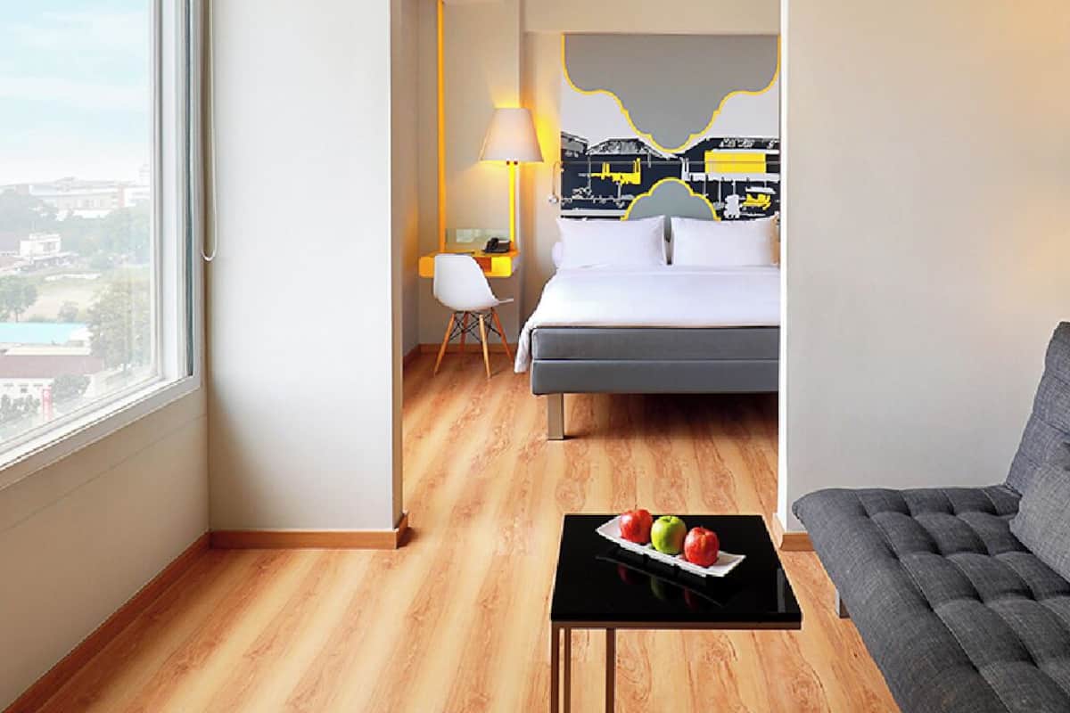 A hotel room with a bed, a desk, a chair, a table with fruit, and a large window.