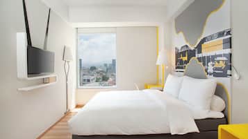 Quarto de hotel moderno com uma cama grande, uma TV fixada na parede, mesa de cabeceira com abajur e vista para a cidade pela janela.