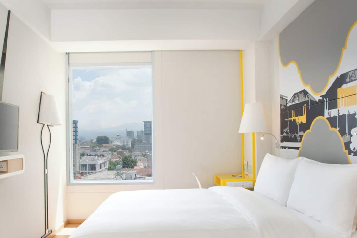 A modern hotel room with a large bed, a city view, a desk with a lamp, and a painting on the wall.