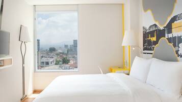 Habitación de hotel moderna con una cama grande, vistas a la ciudad, un escritorio con lámpara y un cuadro en la pared.